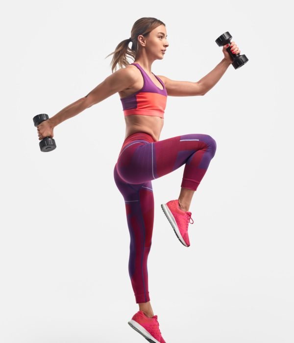 Full body view of muscular young female in sportswear swinging arms with heavy dumbbells and leaping against white background during training for biceps and legs